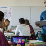 Education Color Photo of K12 class 6th-9th with back table of two females and male with books and then one closer table with one male with windows screen and one female and male teacher talking
