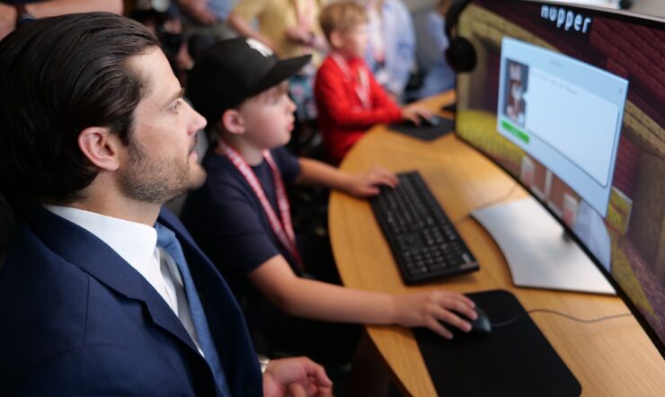 A person in a suit sits beside a child using a computer, with several other people working at computers in the background.