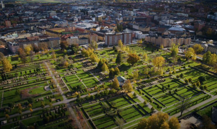 Flygbild över en stor kyrkogård med gröna gräsmattor, gångvägar, gravplatser och träd. Tät stadsbebyggelse syns i bakgrunden.