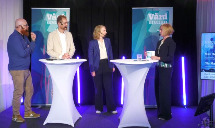 Fyra personer står på en scen och samtalar vid två höga, vita ståbord. I bakgrunden syns två skyltar med texten ”Vårdarenan”, svarta draperier och vertikala ljuslister i blått och lila. Till höger syns även en skärm med text och namn.